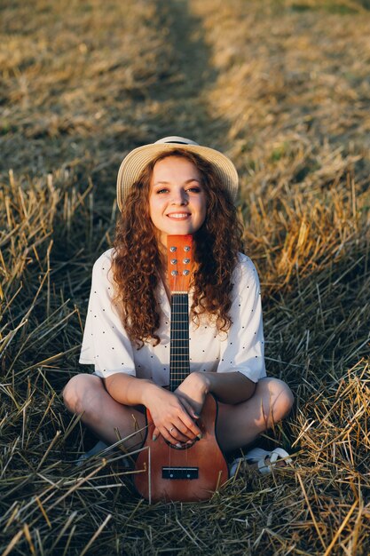 Jonge mooie vrouw met lang krullend haar in korte broek, een hoed met een gitaar vormt in een tarweveld in de zomer bij zonsondergang