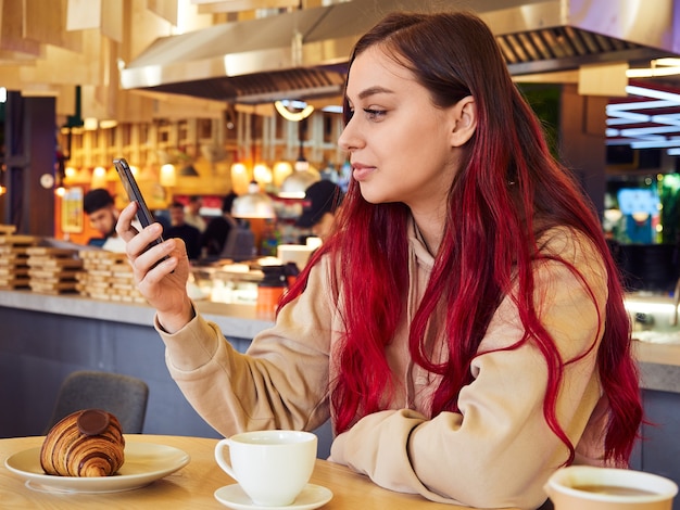 Jonge mooie vrouw met lang rood haar maakt gebruik van een mobiele telefoon in het interieur van een restaurant met een kopje koffie en een croissant