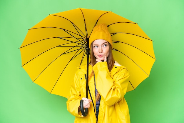 Jonge mooie vrouw met regenbestendige jas en paraplu over geïsoleerde chroma key achtergrond met twijfels en denken