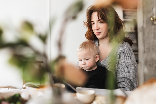 Jonge mooie vrouw met rood haar in gebreide trui zittend aan de tafel dromerig voeden haar zoontje