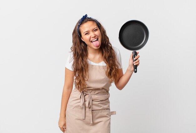 Jonge mooie vrouw met vrolijke en rebelse houding, grappen makend en tong uitsteken chef-kok concept en een pan vasthouden