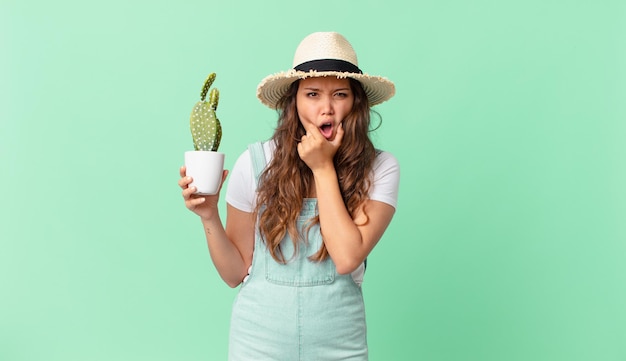 Jonge mooie vrouw met wijd open mond en ogen en hand op kin en met een cactus