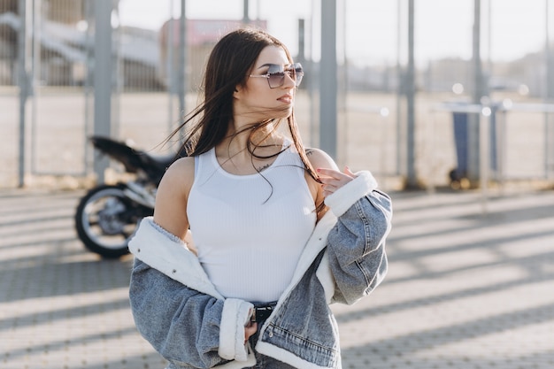 Jonge mooie vrouw met zwart haar in denim broek en een jas in de buurt van een zwarte sportmotorfiets