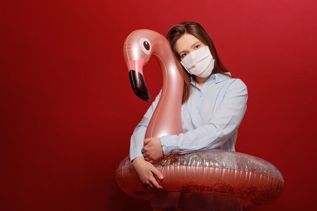 Jonge mooie vrouw op rode muur in beschermend medisch maskeromhelzingen die cirkel roze flamingo zwemmen