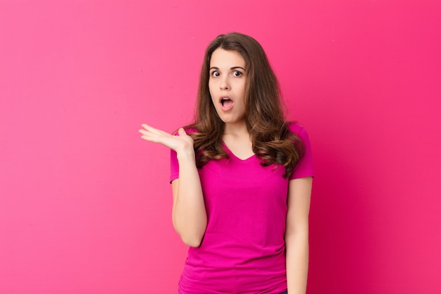 Jonge mooie vrouw op zoek verrast en geschokt, met kaak gevallen met een object met een open hand aan de zijkant over roze muur