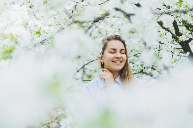 Jonge mooie vrouw staat graag in de buurt van bloeiende lenteboom Een meisje met een beige hoed en een witte jurk glimlacht tussen bloeiende appelbomen Lenteseizoenconcept