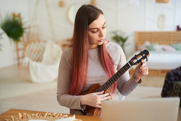 Jonge mooie vrouw stemt ukelele af om te oefenen. Leren op afstand een muziekinstrument te bespelen.