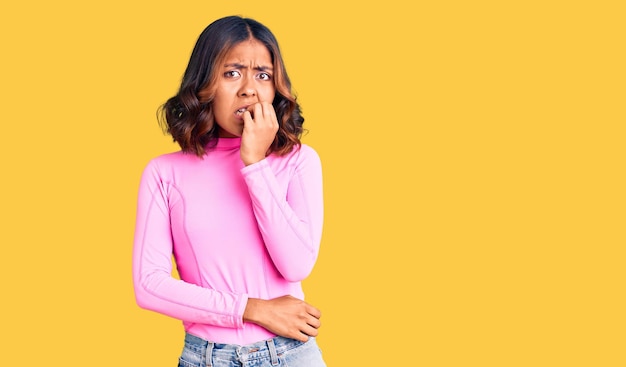 Jonge, mooie vrouw van gemengd ras, gekleed in een roze shirt, ziet er gestrest en nerveus uit met handen op de mond terwijl ze nagels bijt. angst probleem.