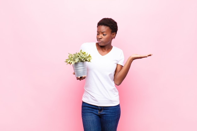 Jonge mooie vrouw verward en verward voelen, twijfelen, wegen of verschillende opties kiezen met grappige uitdrukking die een plant houdt