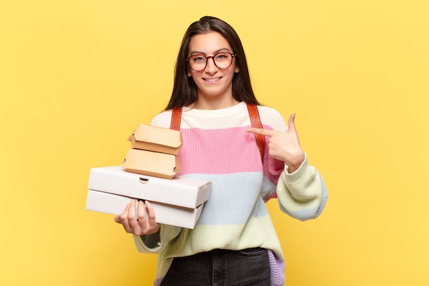 Jonge mooie vrouw voelt zich gelukkig, verrast en trots, wijzend naar zichzelf met een opgewonden, verbaasde blik. neem een fastfoodconcept