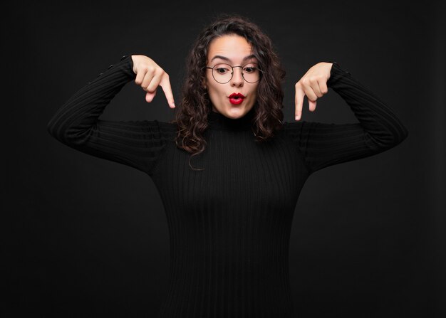 Foto jonge mooie vrouw voelt zich geschokt, met open mond en verbaasd, kijkt en wijst naar beneden in ongeloof en verrassing over zwarte muur.