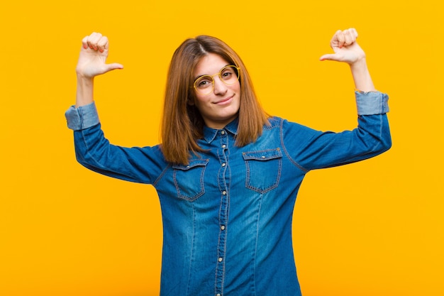 Jonge mooie vrouw voelt zich trots, arrogant en zelfverzekerd, kijkt tevreden en succesvol, wijzend naar zichzelf tegen gele muur