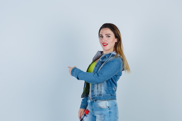 Jonge mooie vrouw wijst terug in denim outfit en ziet er vrolijk uit, vooraanzicht.