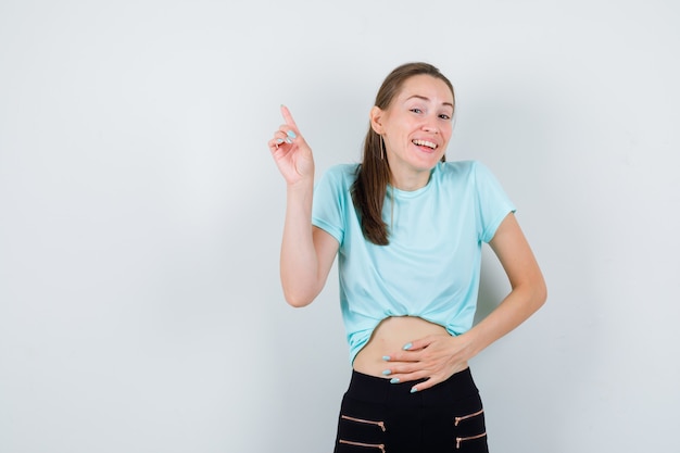 Jonge mooie vrouw wijzend naar de linker bovenhoek, met de hand op de buik in t-shirt, broek en blij kijken. vooraanzicht.