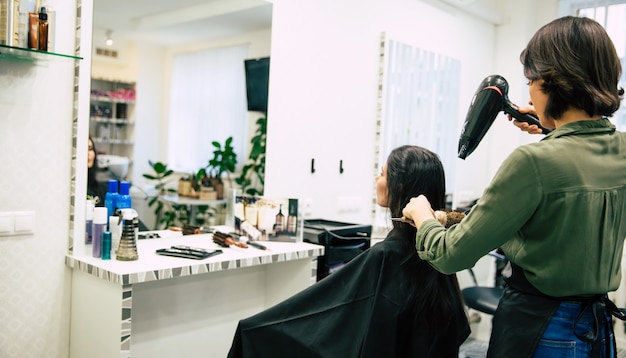 Jonge mooie vrouw zit in de fauteuil in een schoonheidssalon en laat haar haar drogen door een lachende professionele stylist.