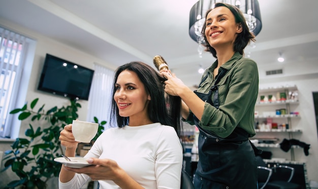 Jonge mooie vrouw zit in de fauteuil in een schoonheidssalon en laat haar haar drogen door een lachende professionele stylist.