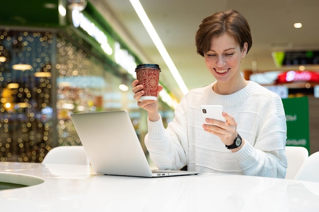 Jonge mooie vrouw zit in een winkelcentrum aan een tafel en werkt op een computerlaptop met behulp van