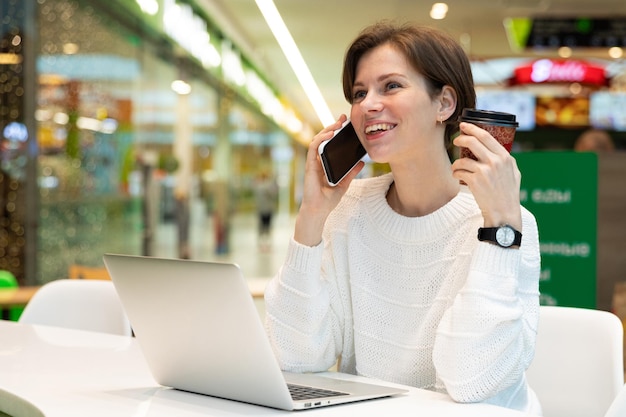 Jonge mooie vrouw zit in een winkelcentrum aan een tafel en werkt op een computerlaptop met behulp van