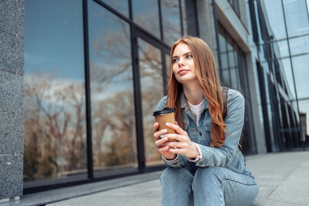 Jonge mooie vrouw zit op de trap in de buurt van het zakencentrum en drinkt koffie