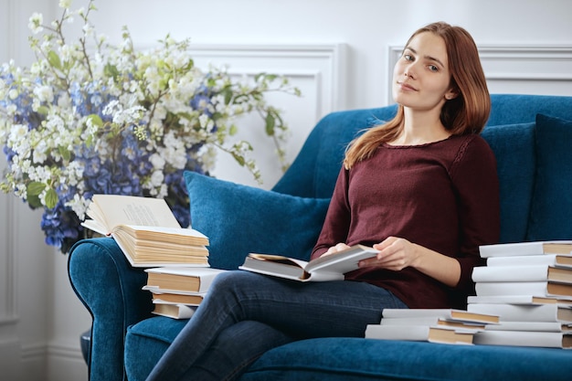 Jonge mooie vrouw zit op een bank tussen een stapel boeken