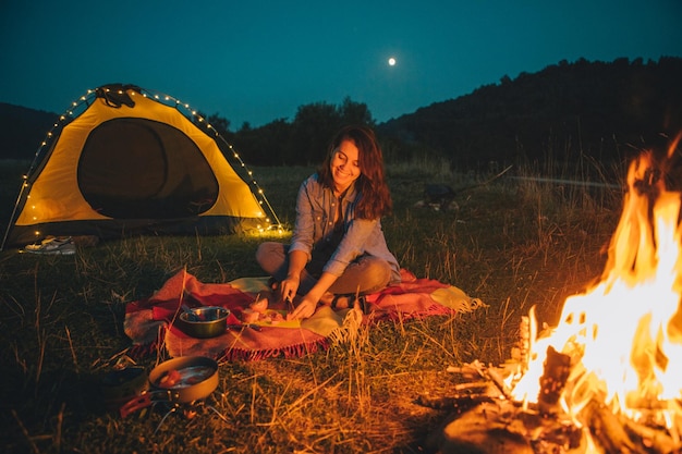 Jonge mooie vrouw zittend op de grond voor vuur koken diner gele tent op achtergrond