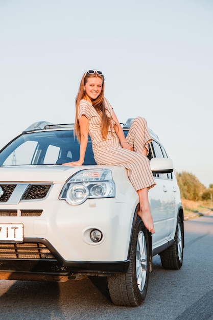 Jonge mooie vrouw zittend op de motorkap van de auto bij zonsondergang