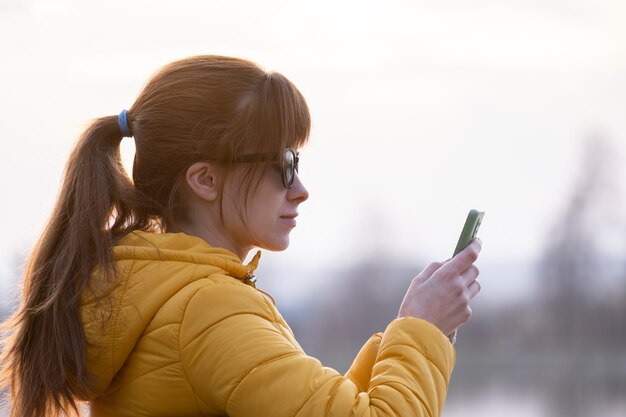 Jonge mooie vrouw zittend op een bankje in het park bladeren door haar smartphone buiten op warme herfstdag.