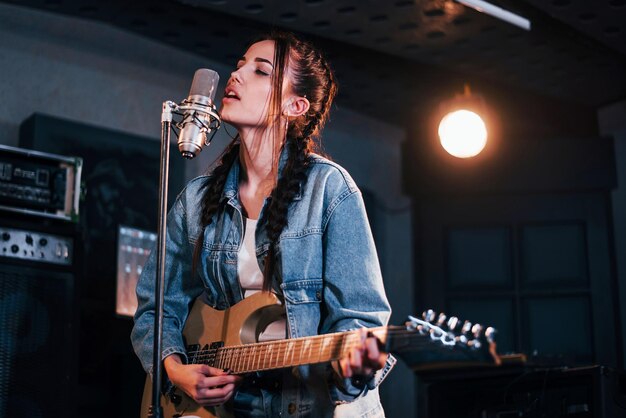 Jonge mooie vrouwelijke artiest met gitaar zingen en repeteren in een opnamestudio