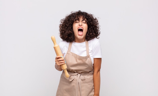 Jonge mooie vrouwelijke chef-kok die agressief schreeuwt, er erg boos, gefrustreerd, verontwaardigd of geïrriteerd uitziet, nee schreeuwt