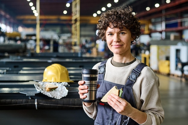 Jonge mooie vrouwelijke ingenieur met kopje thee en sandwich camera kijken