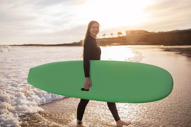 Foto jonge mooie vrouwelijke surfer met surfplank poseren op strand bij zonsondergang