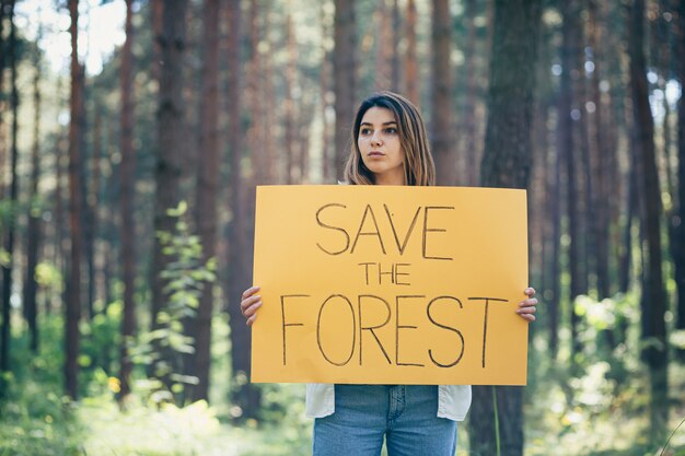 Jonge, mooie vrouwelijke vrijwilligersactivist in het bos met een poster, red het bos