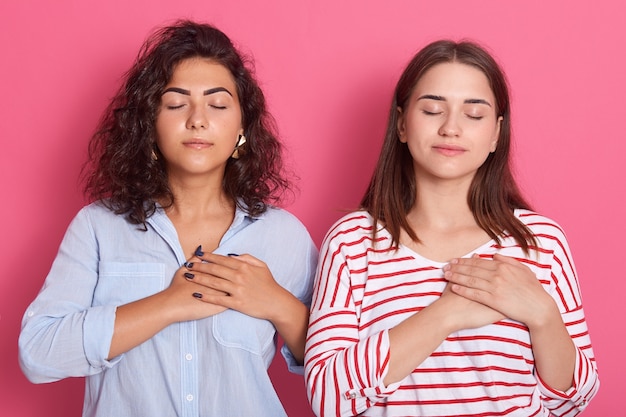 Jonge mooie vrouwen die vrijetijdskleding dragen die zich geïsoleerd over roze achtergrond met handen op borst bevinden