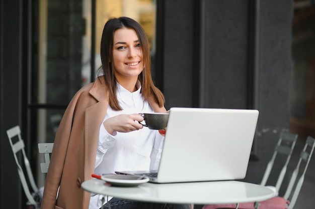 Jonge mooie zakenvrouw die op laptop in straatcafé werkt, typt op toetsenbord, slimme dame die serieus op het scherm kijkt. Smartphone en bril op tafel. Het dragen van stijlvolle roze jas, witte horloges.