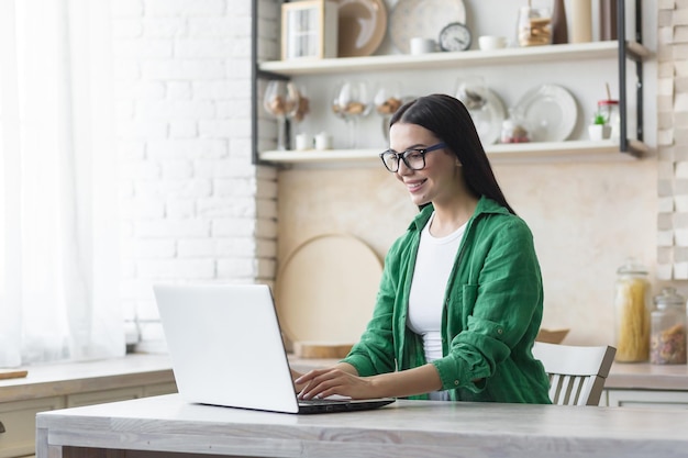 Jonge mooie zakenvrouw in glazen en groen shirt thuis werken in de keuken op laptop