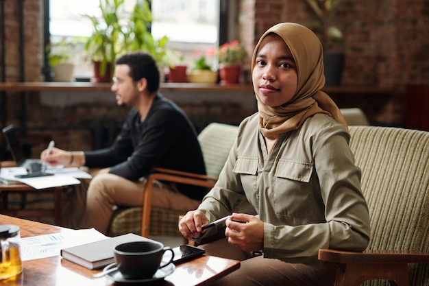 Jonge mooie zakenvrouw in hijab aan tafel zitten en kijken naar de camera