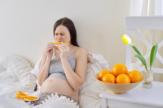 Jonge mooie zwangere vrouw die sinaasappelen eet die thuis in bed zitten. Gezonde voeding, natuurlijke vitamine C