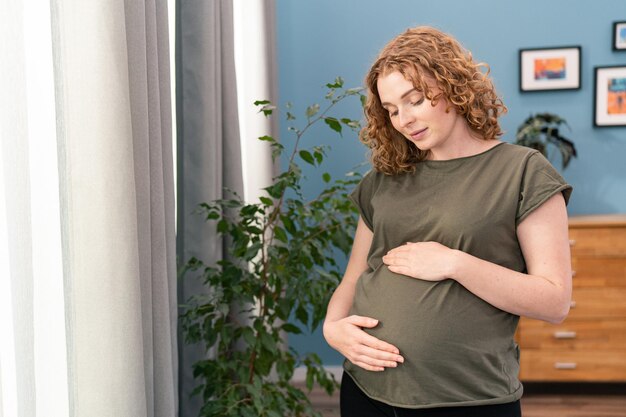 Jonge mooie zwangere vrouw die thuis bij het raam staat