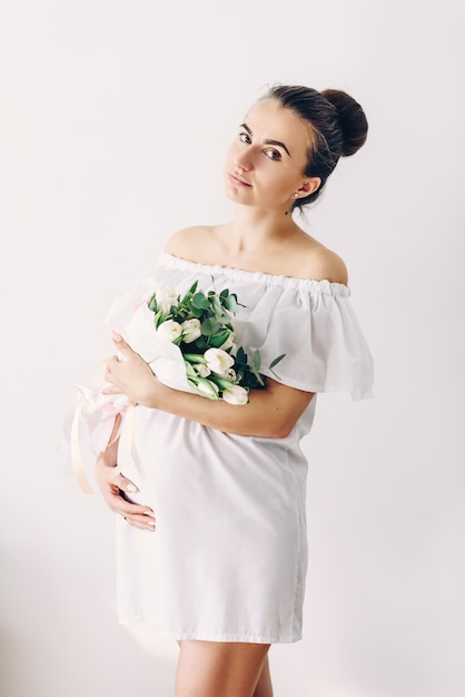 Jonge mooie zwangere vrouw in een witte jurk met een boeket van witte tulpen