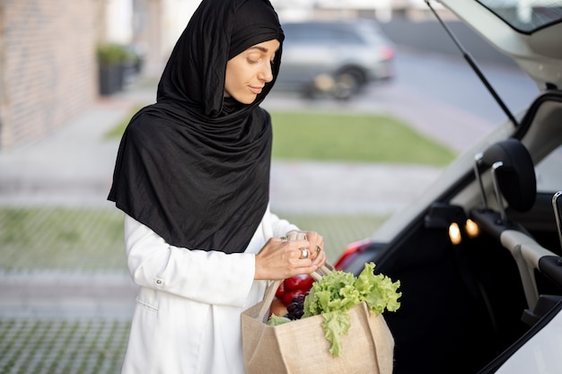 Jonge moslim zakenvrouw in hijab naar huis met boodschappen, staande met boodschappentas op de veranda van haar huis