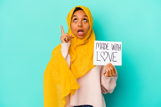 Jonge moslimvrouw die een gemaakt met liefdeaanplakbiljet houdt dat op blauwe muur wordt geïsoleerd die met geopende mond ondersteboven richt.