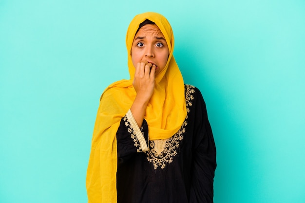 Jonge moslimvrouw die op blauwe muur vingernagels bijt, zenuwachtig en zeer angstig