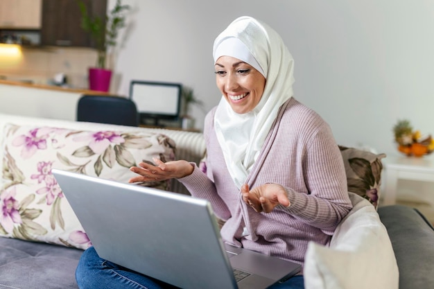 Jonge moslimvrouw die thuis een videogesprek via laptop voert Gelukkige glimlachende moslimvrouw zit op de bank en gebruikt laptop in de woonkamer thuis om taalvideo's te leren bellen