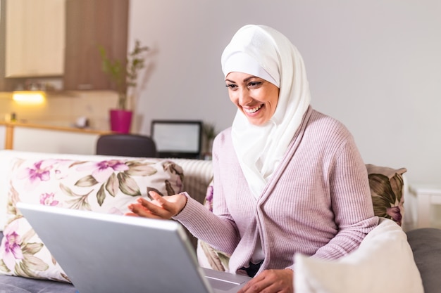 Jonge moslimvrouw die videogesprek via laptop thuis. Gelukkig lachend moslimvrouw zittend op de bank