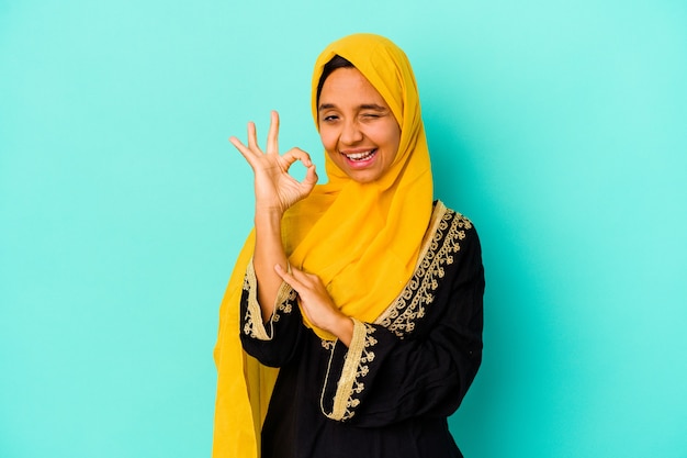 Jonge moslimvrouw geïsoleerd op blauwe achtergrond knipoogt en houdt een goed gebaar met de hand.