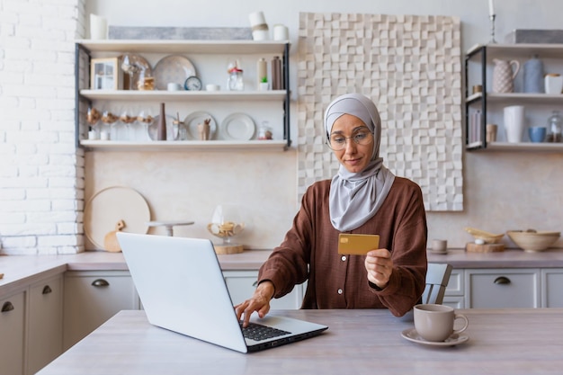 Jonge moslimvrouw in hijab die op afstand online werkt terwijl hij in de keuken zit te typen op een laptop en