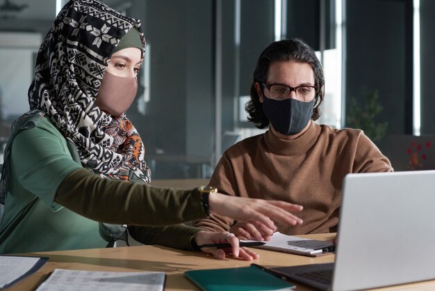 Jonge moslimvrouw in masker die online presentatie met haar partner bespreekt tijdens vergadering op kantoor