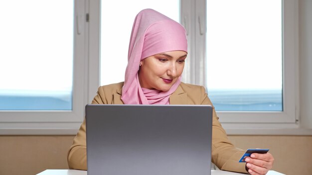 Jonge moslimvrouw met een roze hoofddoek doet online aankopen op een laptop met een bankkaart in handen.