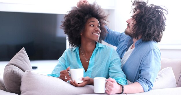 jonge multi-etnische paar zittend op de bank thuis koffie drinken, praten, glimlachen.