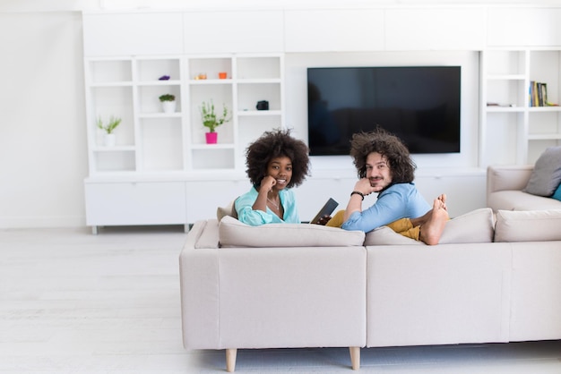 Jonge multi-etnische paarzitting op een bank in de luxewoonkamer, die een tabletcomputer met behulp van